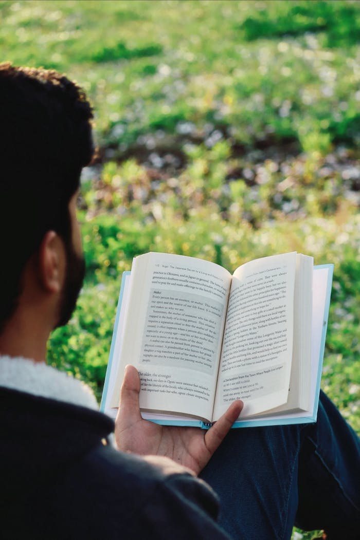 A man with facial hair sits outdoors, reading a book amidst greenery on a sunny day.